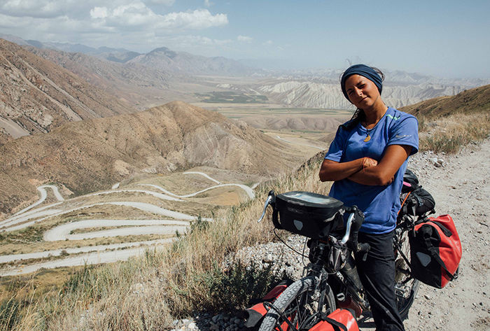 Nicole poses with her arms crossed, standing on a high ledge with a winding mountain road in the background