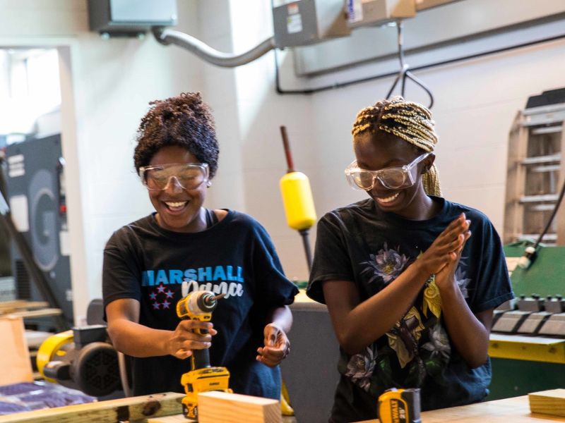 Two smiling students wearing protective goggles handle power tools and lumber