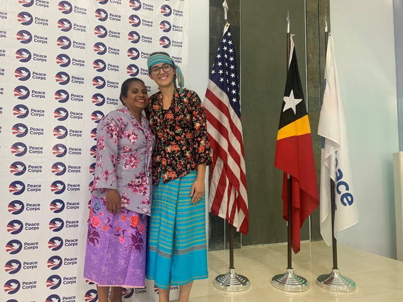 Two people smile, arm-in-arm, wearing colorful garb in front of a Peace Corps banner