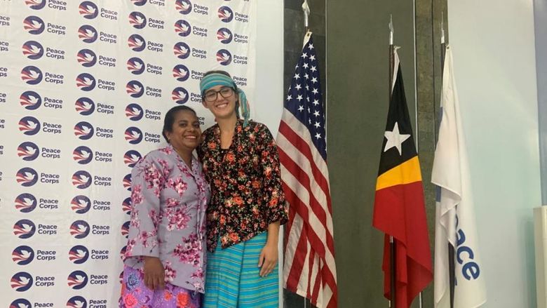 Two people smile, arm-in-arm, wearing colorful garb in front of a Peace Corps banner