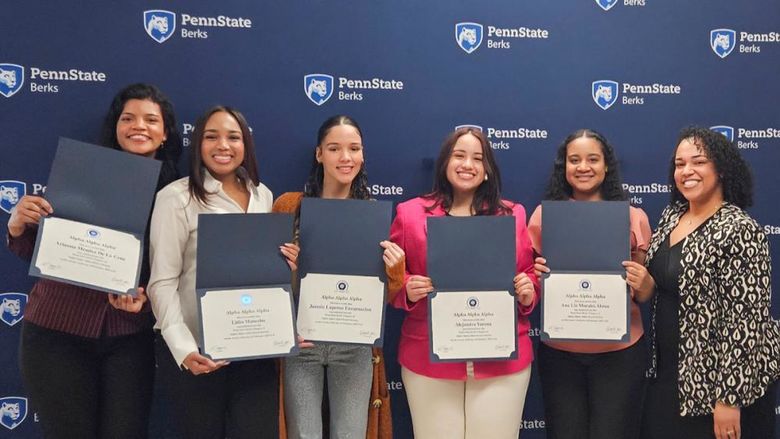 Five students hold certificates open with a staff member, all standing in front of a branded Penn State banner