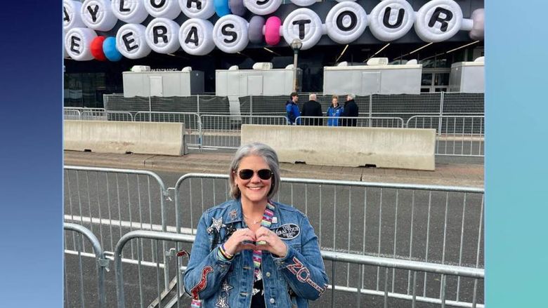 Professor stands in front of marquee reading "Taylor Swift Eras Tour" while making a heart with her hands