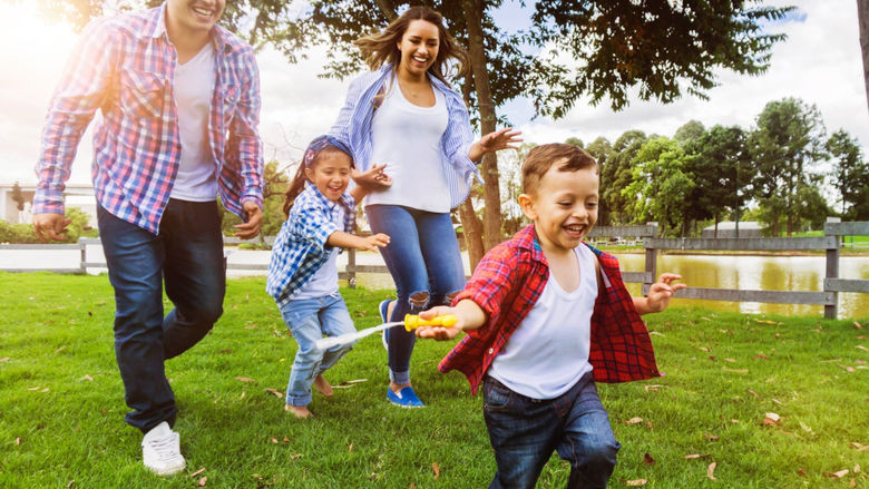 A man, a woman, and two children smile as they run through a park. trees and a fence are in the background.