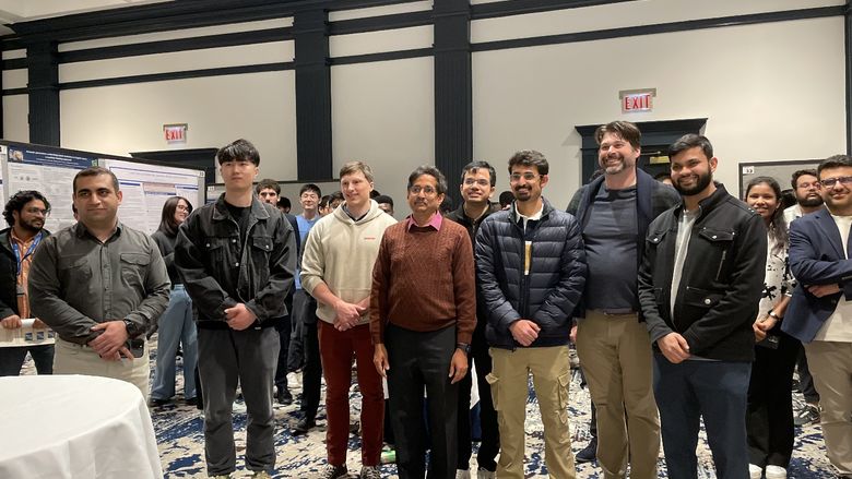Penn State students and faculty stand in a line for a photo surrounded by their research posters during poster session.