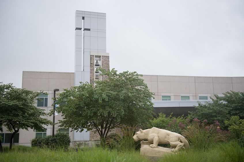 A statue of the Penn State Nittany Lion at the Penn State Health St. Joseph campus appears in profile in front of a row of trees and weeds. Behind the scene rises a glass tower and bells and windows.
