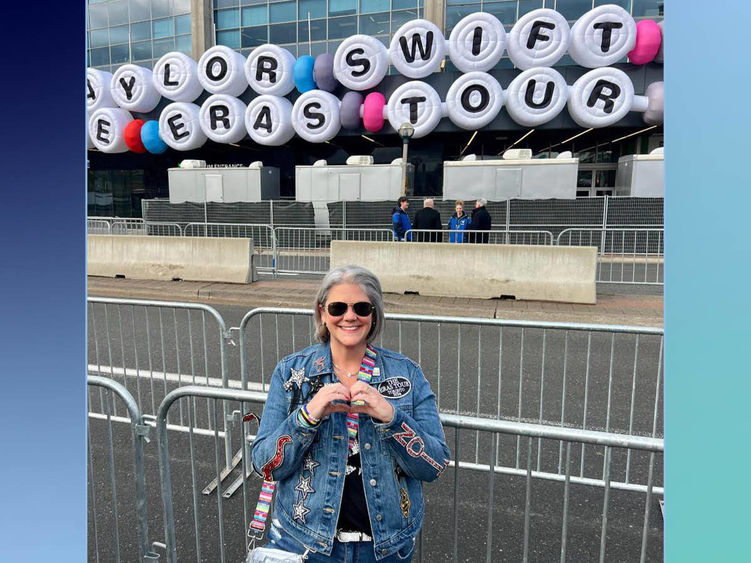 Professor stands in front of marquee reading "Taylor Swift Eras Tour" while making a heart with her hands