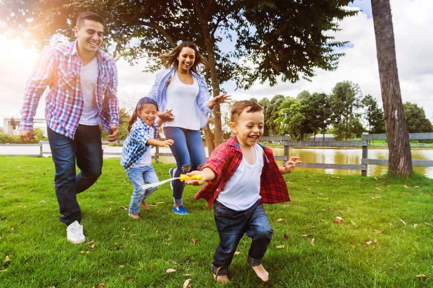 A man, a woman, and two children smile as they run through a park. trees and a fence are in the background.